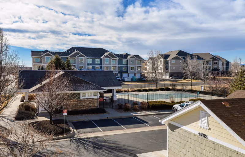 View of a senior living facility with multiple residential buildings, a fenced outdoor pool area, parking spaces, and leafless trees under a partly cloudy sky.