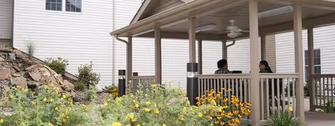 A covered outdoor gazebo with two people seated inside, surrounded by flowering plants, landscaping rocks, and the side of the building.