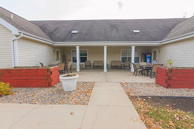 Covered outdoor patio of a single-story assisted living building with chairs, tables, planters, and a vending machine.