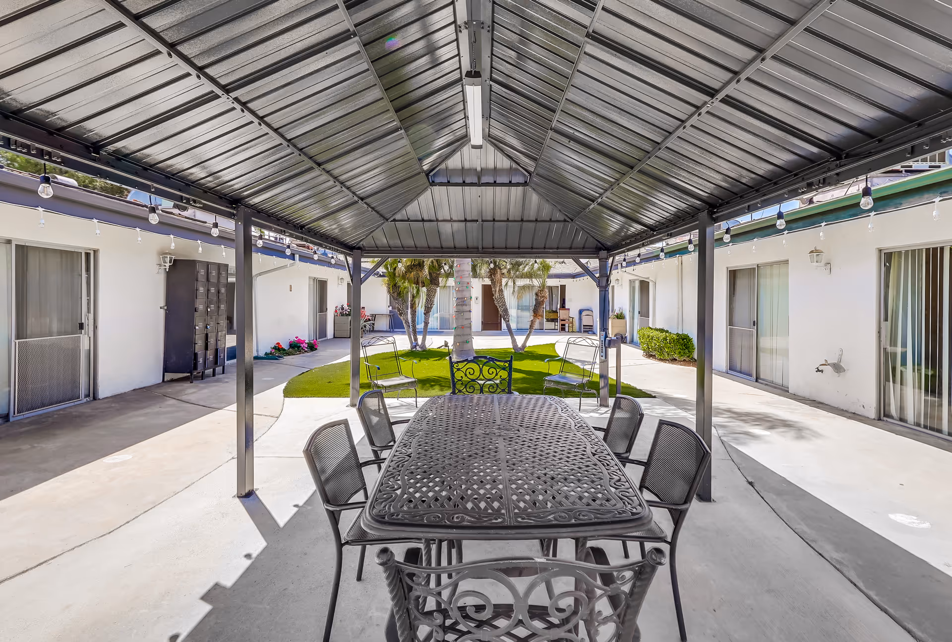 Outdoor covered patio area with a metal roof and a long metal table surrounded by six chairs. The patio is part of a courtyard with a green lawn, palm trees, and white buildings with sliding glass doors surrounding the space.