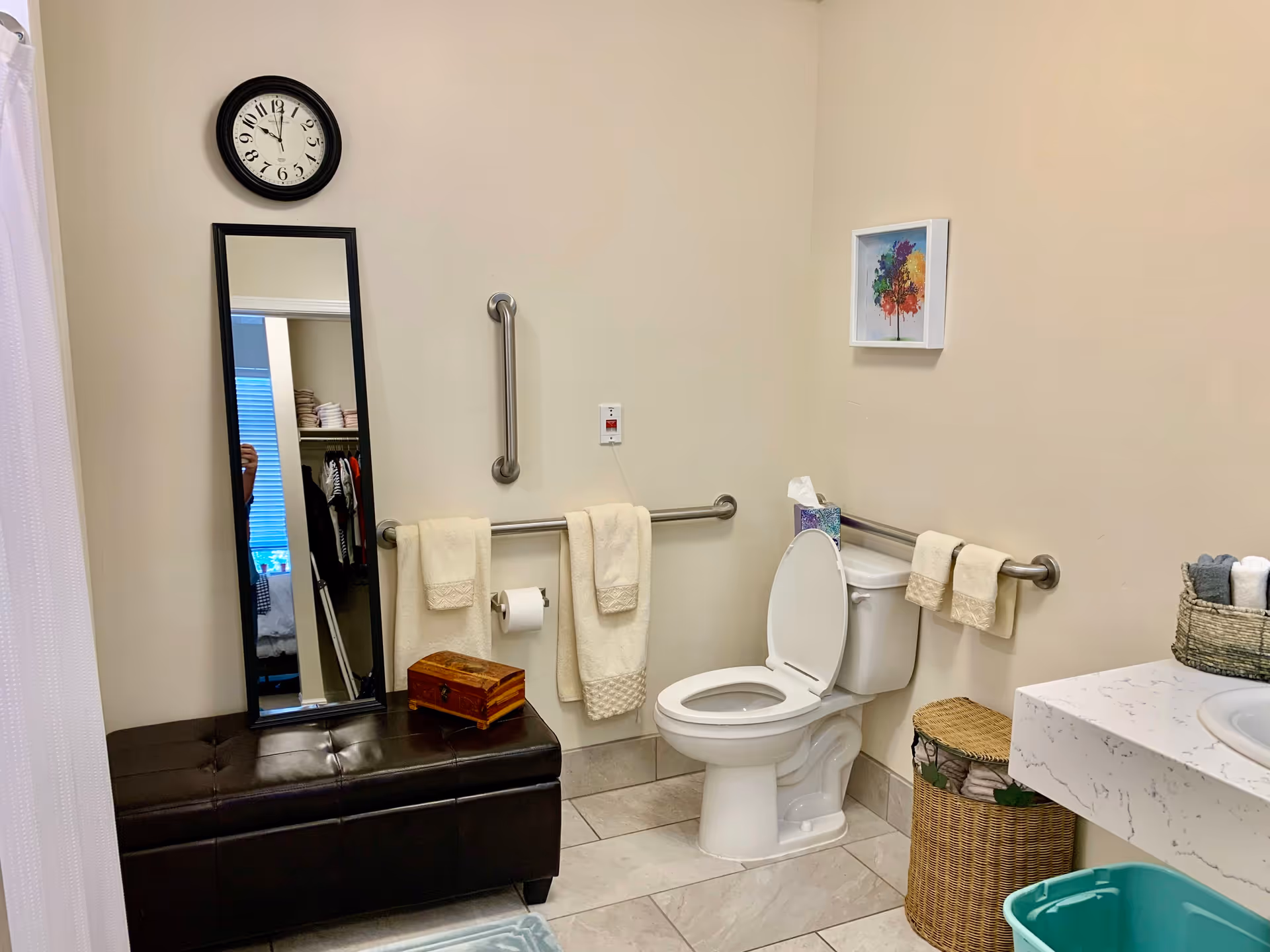 A clean and accessible bathroom with a white toilet, grab bars on the walls, beige towels hanging on the bars, a wicker basket, a black cushioned bench with a wooden box on top, a tall mirror leaning against the wall, a clock above the mirror, and a colorful framed picture on the wall.