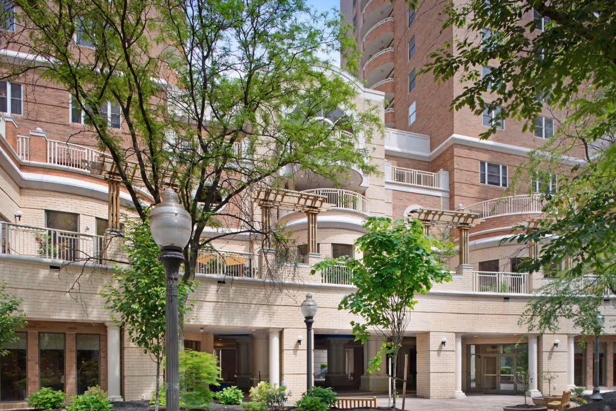 Exterior courtyard and facade of a multi-story brick senior living building with balconies, trees, and lampposts.