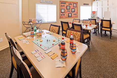 Light-filled communal dining/activity room with tables arranged for a board game, colorful cups and bottled drinks.