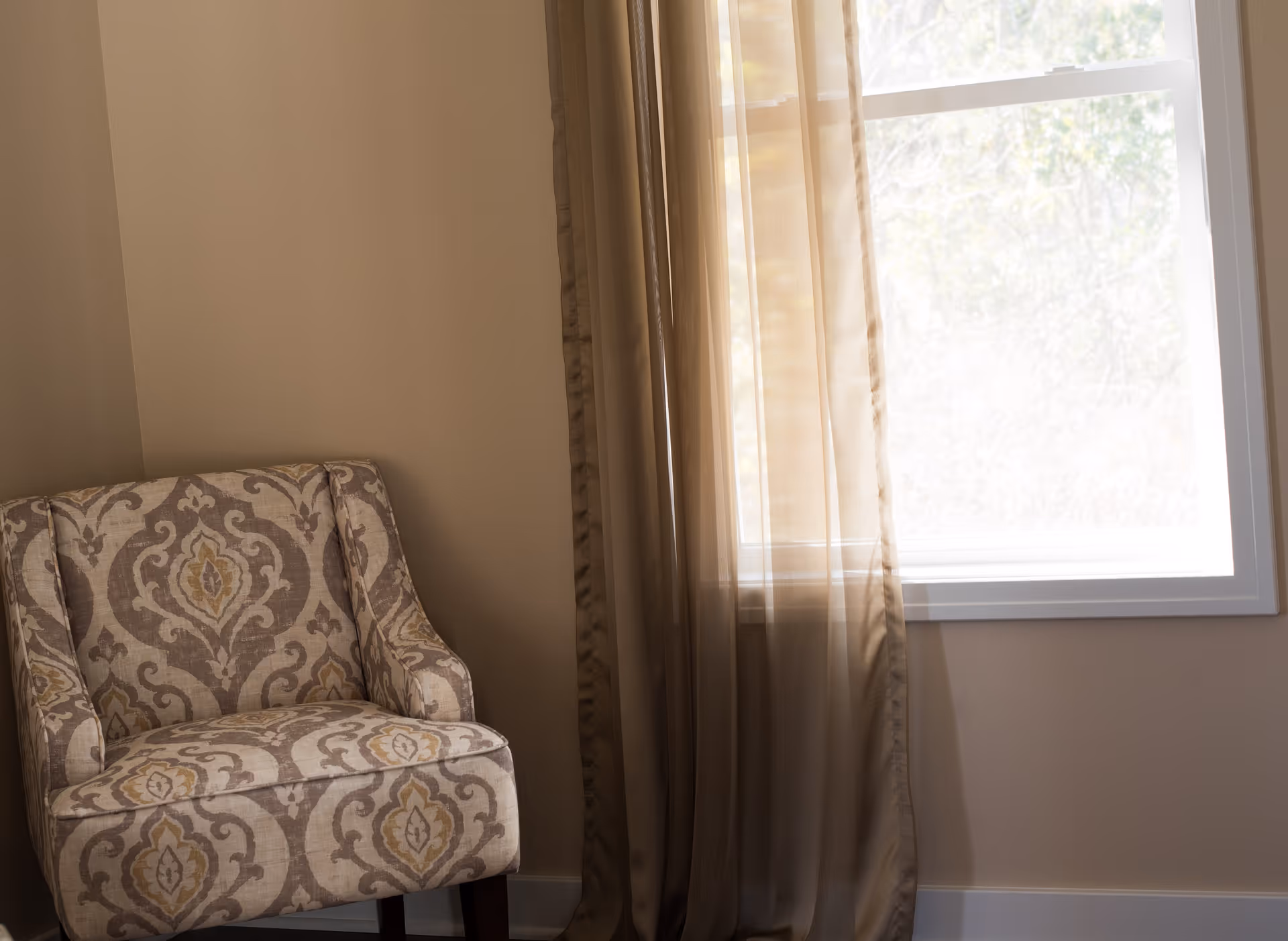 A patterned upholstered armchair next to a window with sheer beige curtains, in a softly lit room with beige walls.