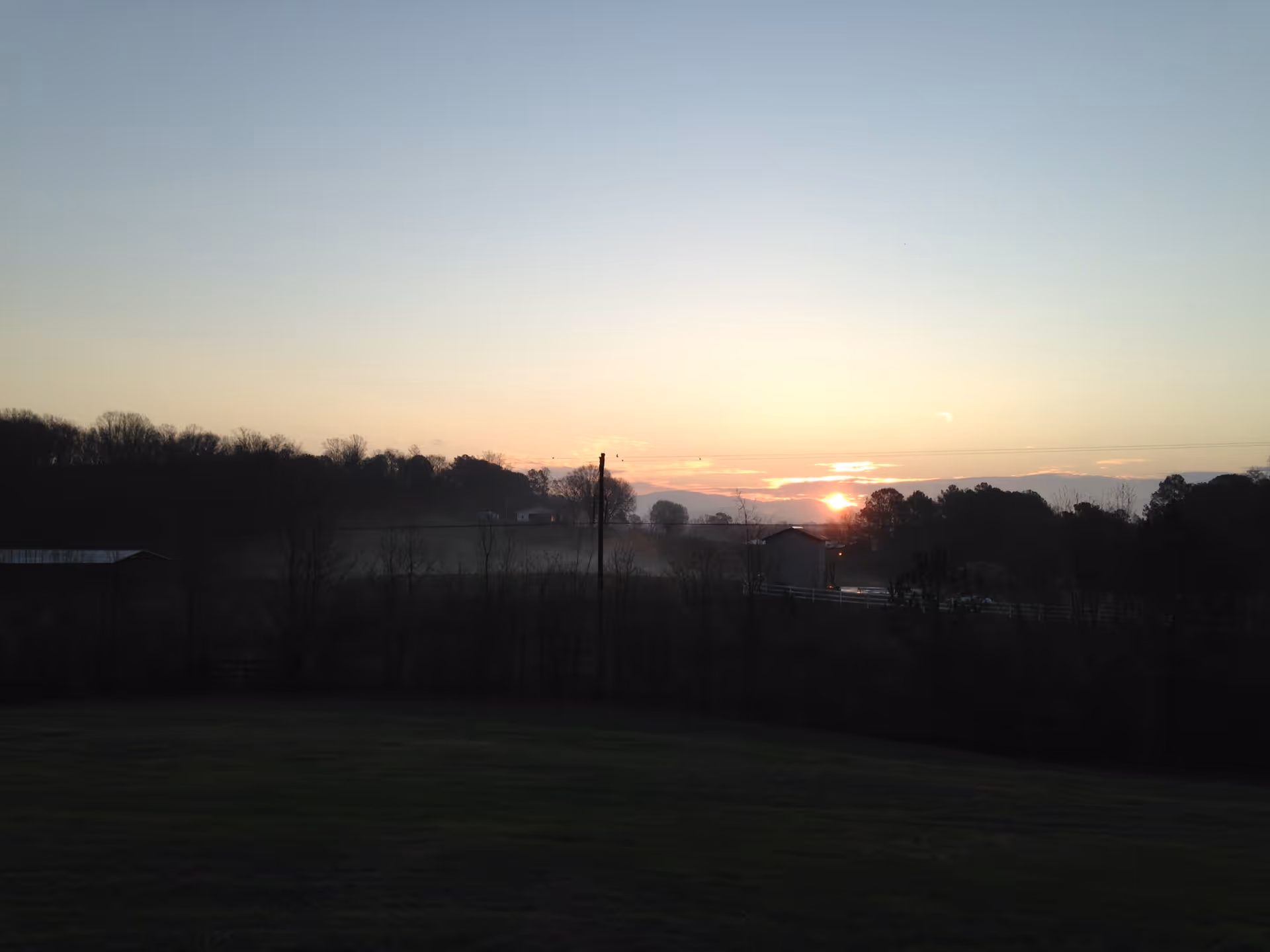 A rural landscape at sunset with a grassy field in the foreground, leafless trees, a few buildings, and hills in the background under a clear sky.