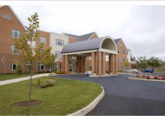 Front exterior view of a multi-story brick building with a covered entrance supported by four brick columns. There is a driveway leading up to the entrance, landscaped grass areas with small trees and flower beds, and a few parked cars visible in the background under a cloudy sky.