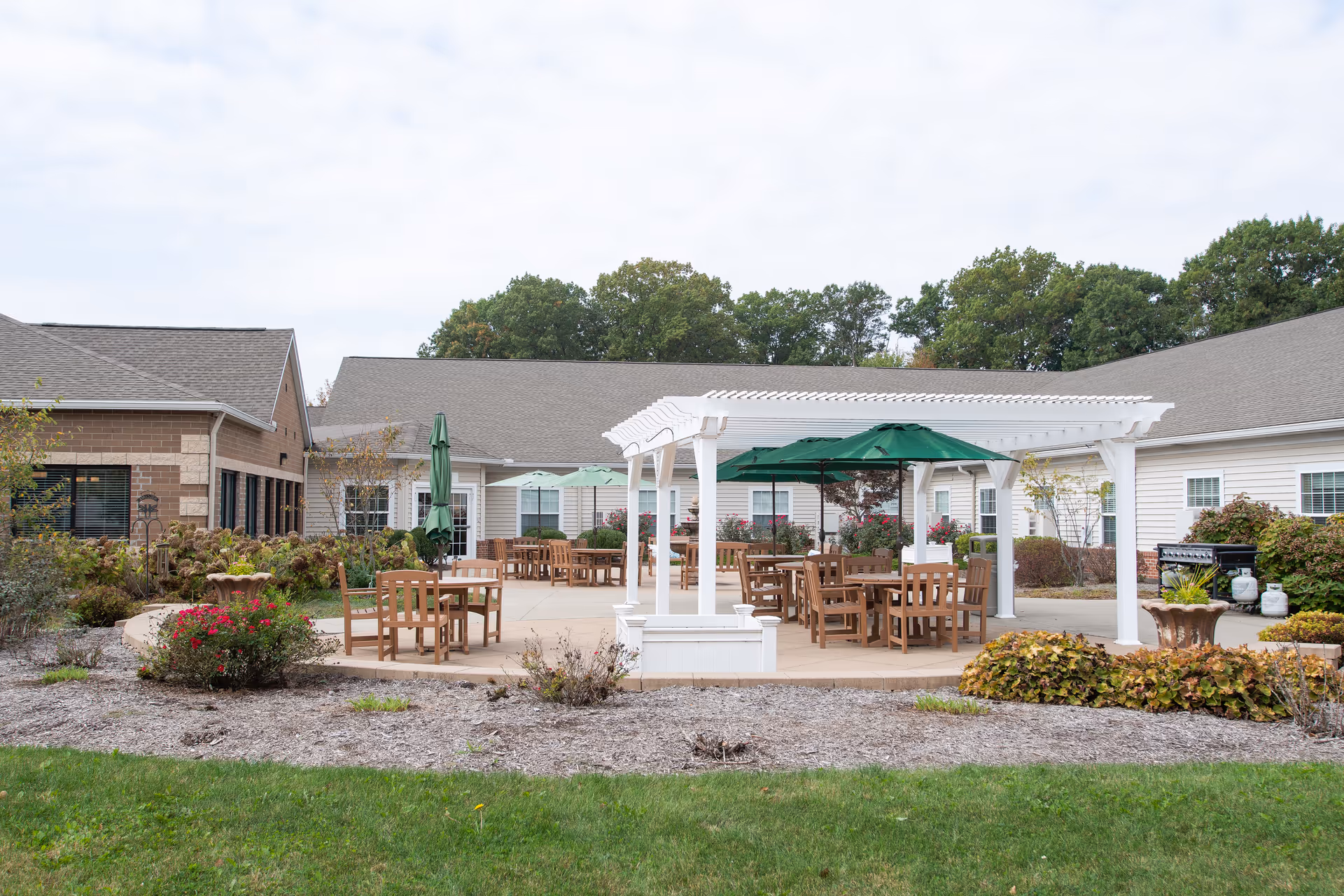 Outdoor patio area at Danbury Massillon with wooden tables and chairs, green umbrellas, a white pergola, surrounding bushes and plants, and a barbecue grill near the building. The building has beige siding and a gray roof, with trees in the background under a cloudy sky.