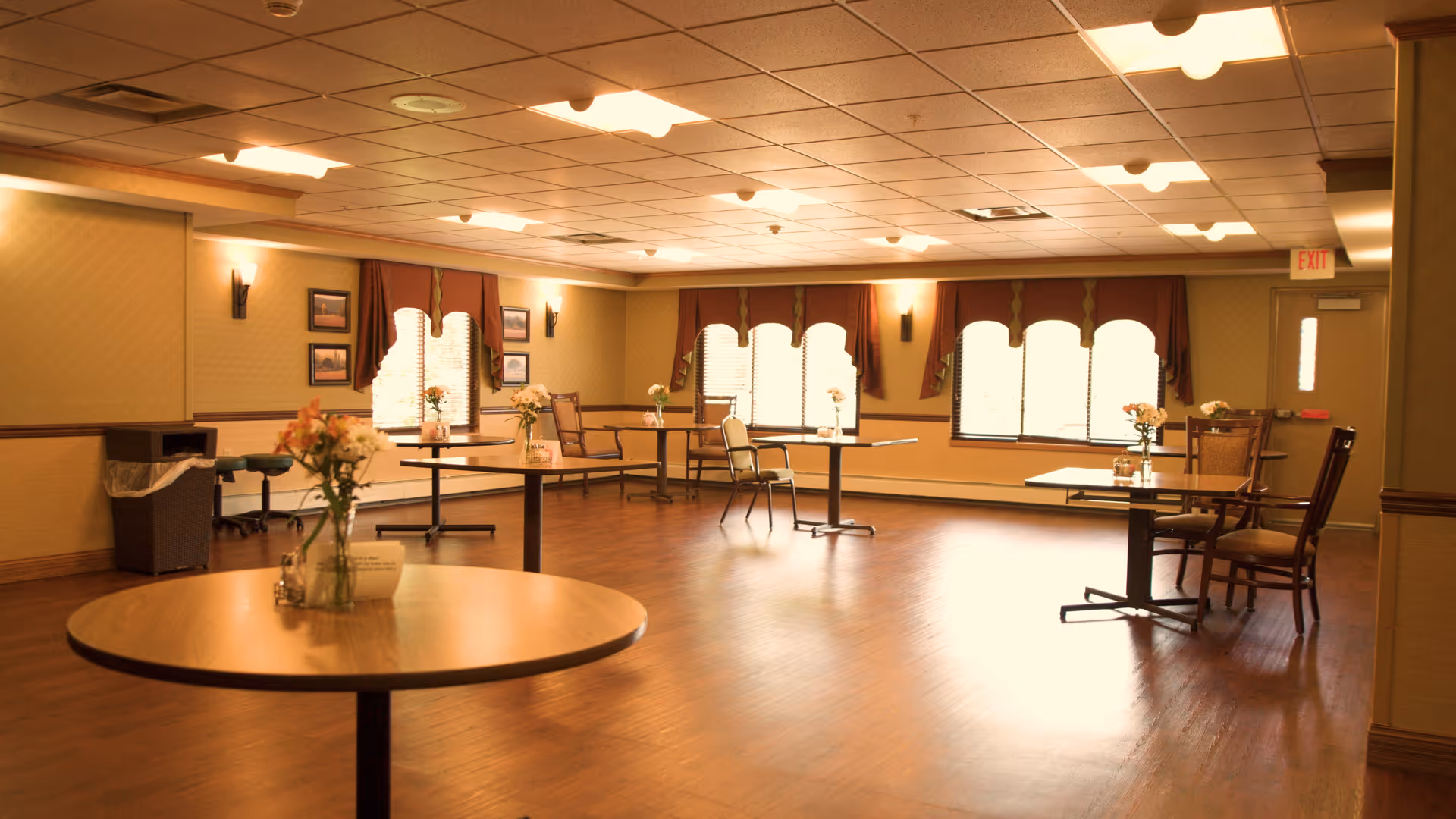 Spacious dining/activity room with round tables, chairs, flower centerpieces, and curtained windows.