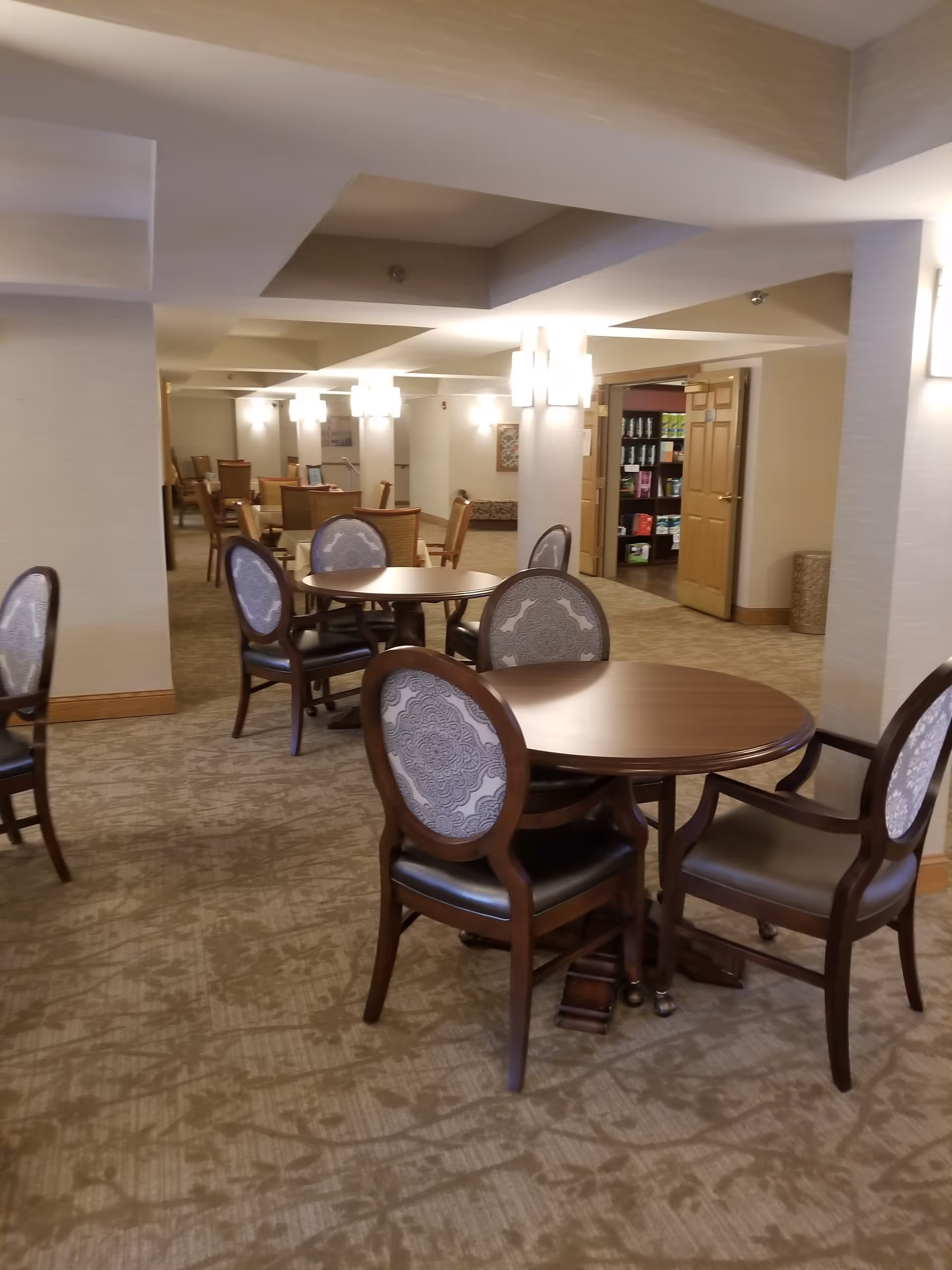 Interior view of a senior living facility common area with round wooden tables and upholstered chairs arranged on a patterned carpet. The room is softly lit with wall sconces and ceiling lights, and there is an open doorway leading to a room with shelves stocked with various items.