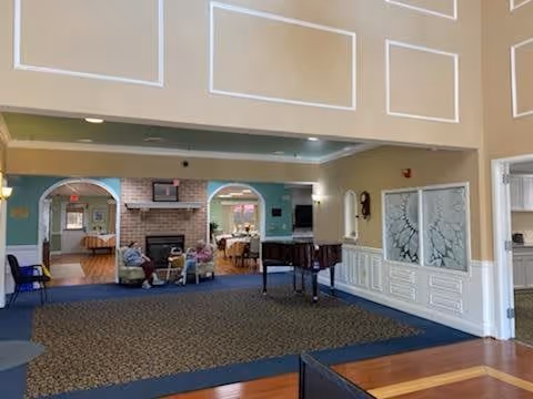 Interior view of a senior living facility common area with a carpeted floor and a piano. In the background, there are two elderly people sitting near a fireplace with a television mounted above it. The walls are painted in light colors with decorative molding, and there are archways leading to other rooms.