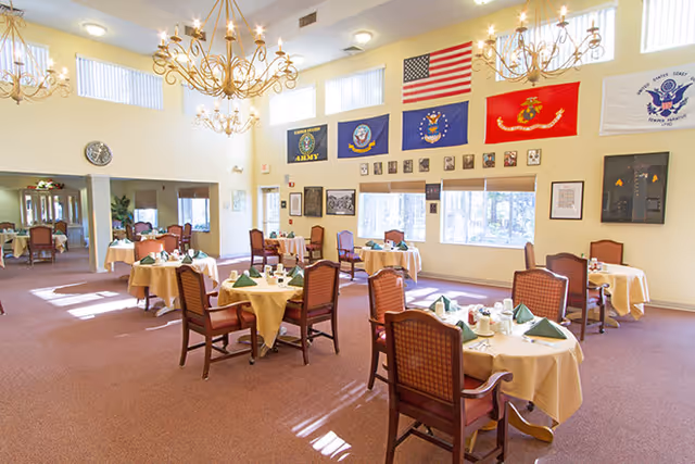 A bright dining room with several round tables covered with beige tablecloths, each set with green folded napkins, cups, and condiments. The room features high ceilings with large chandeliers, multiple windows letting in natural light, and various military flags and framed pictures displayed on the walls. Wooden chairs with cushioned seats surround the tables.