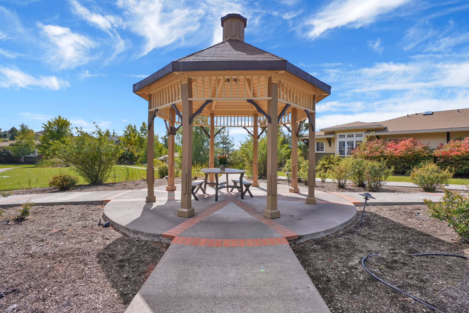 A wooden gazebo with a hexagonal roof stands on a concrete circular platform with a brick border. Inside the gazebo, there is a round table with a flower pot and two benches. The surrounding area features landscaped bushes, a green lawn, and a building with large windows under a blue sky with scattered clouds.