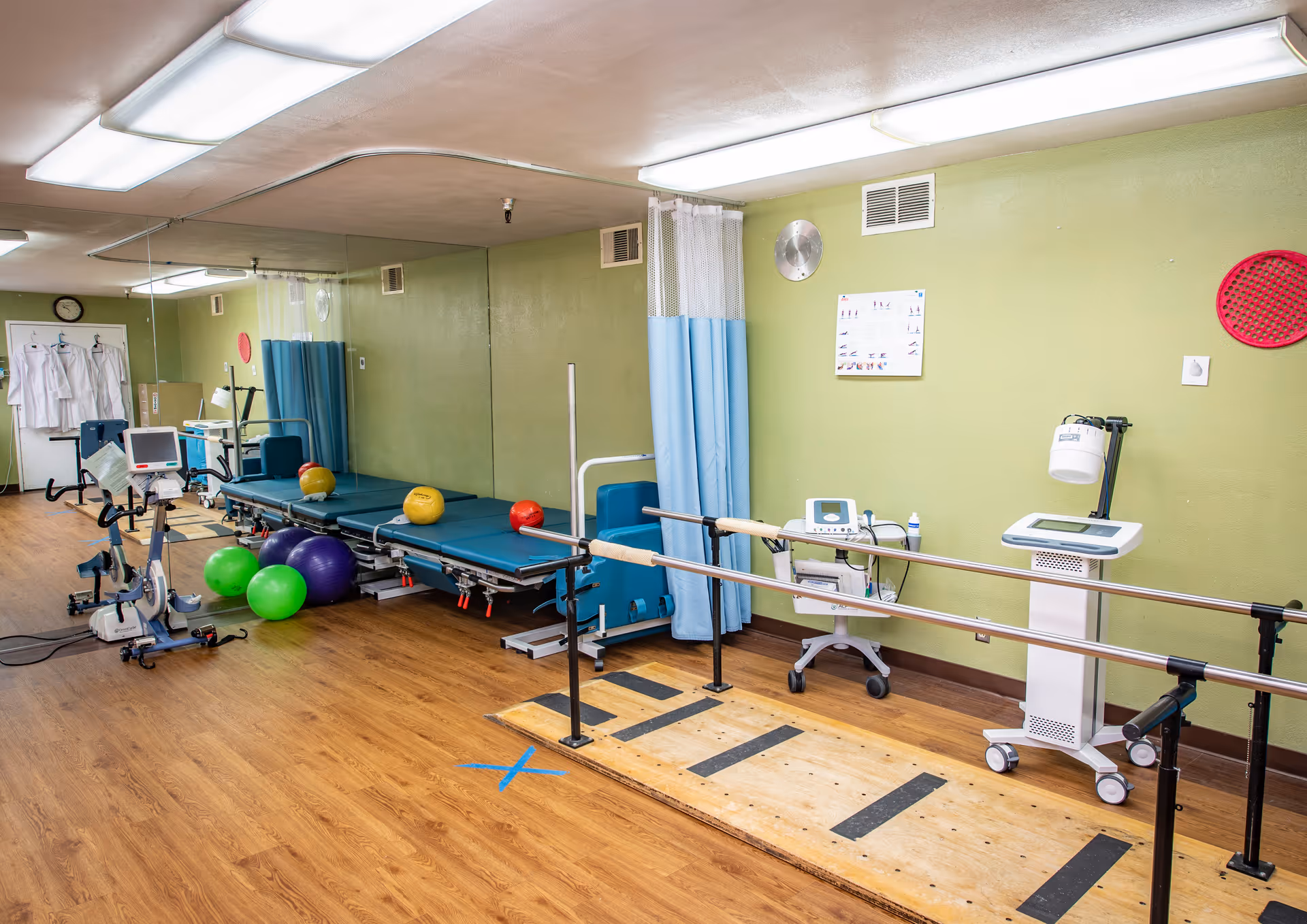 Physical therapy room with exercise equipment including stationary bikes, therapy beds with colorful exercise balls, parallel bars for walking exercises, and medical devices against a green wall with a wooden floor.