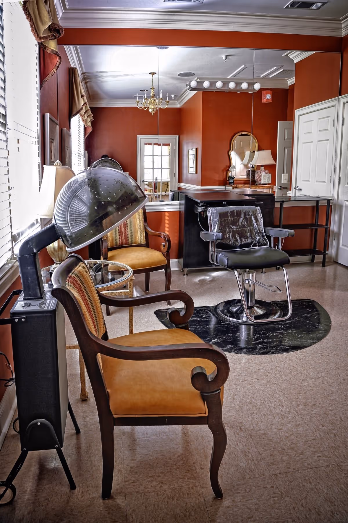 Interior of a senior care facility room with two upholstered chairs, a hair dryer chair with a hooded hair dryer, a large mirror on the wall, a chandelier, and a small table with a lamp. The walls are painted a warm reddish-brown color.