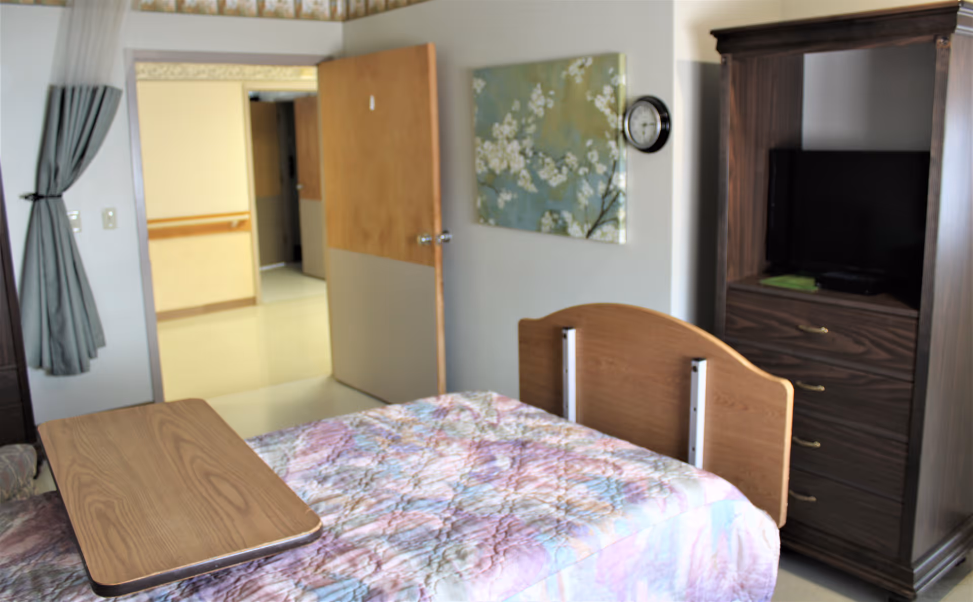 A bedroom in a senior living facility with a bed covered by a pastel-colored quilt. There is a wooden overbed table on the bed, a wooden armoire with a TV inside, a floral painting on the wall, a clock, and an open door leading to a hallway. A window with gray curtains is partially visible on the left.