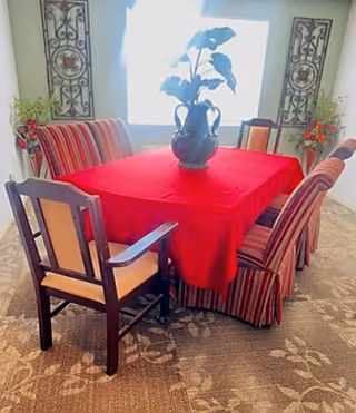 A dining area with a rectangular table covered with a red tablecloth. The table is surrounded by six chairs, four of which have striped upholstery and two have wooden frames with beige cushions. A black vase with green leafy plants is placed in the center of the table. The room has a patterned carpet and two decorative wall hangings on either side of a window in the background.