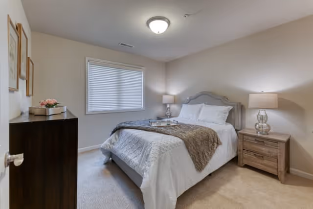 A cozy bedroom with a neatly made bed featuring white bedding and a brown throw blanket. There are two bedside tables with matching lamps on either side of the bed. A window with closed blinds is on the wall behind the bed. A dark wooden dresser with decorative items is positioned near the door. The room has beige walls and carpeted flooring.