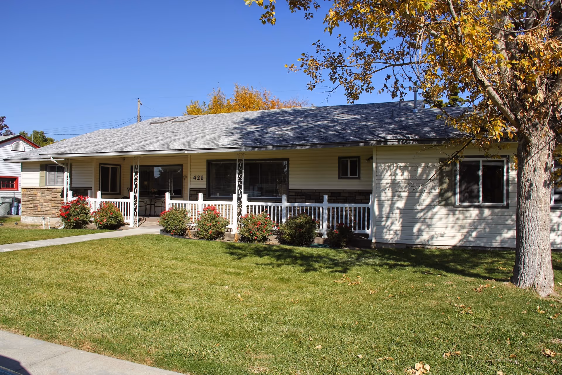 Single-story residential building with beige siding and a gray shingled roof, featuring a white railing porch with several bushes and flowers in front. A large tree with autumn leaves is on the right side of the image, and the lawn is green with a concrete walkway leading to the porch. The building number 421 is visible near the entrance.
