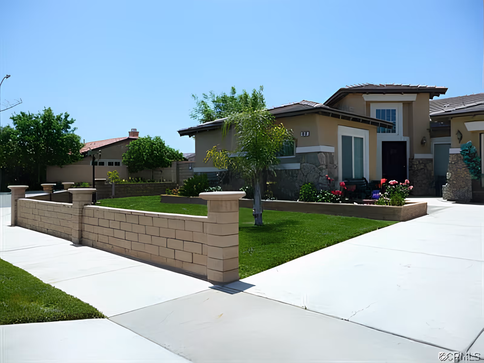 Exterior view of a single-story residential building with beige walls and stone accents, surrounded by a low brick wall and well-maintained green lawn with small trees and flowering plants under a clear blue sky.