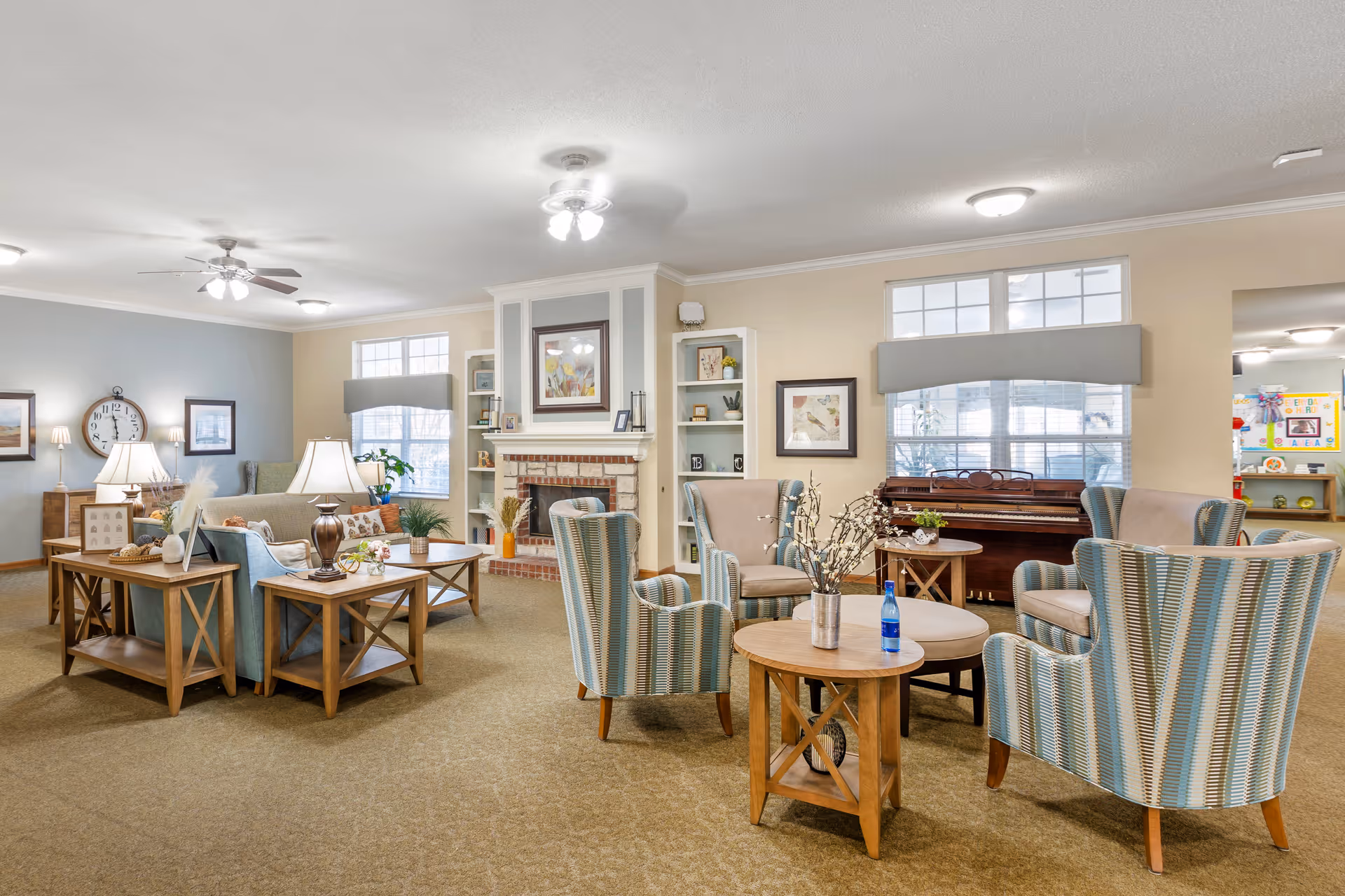 A spacious and well-lit living room area with multiple seating arrangements including armchairs and sofas around wooden coffee tables. The room features a brick fireplace with framed artwork above it, built-in shelves with decorative items, large windows with valances, a piano against the wall, and ceiling fans with lights. The decor is warm and inviting with neutral tones and soft lighting.