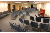 Large interior meeting room with rows of gray chairs arranged facing a small stage holding an upholstered armchair and lectern.