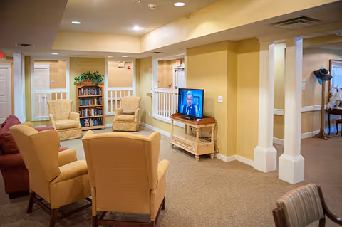 A cozy living room area in a senior living facility with beige walls and carpet. The room features several upholstered armchairs arranged around a small wooden TV stand with a flat-screen TV. There is a bookshelf filled with books against the far wall, and white railings and columns separate the space from adjacent hallways.