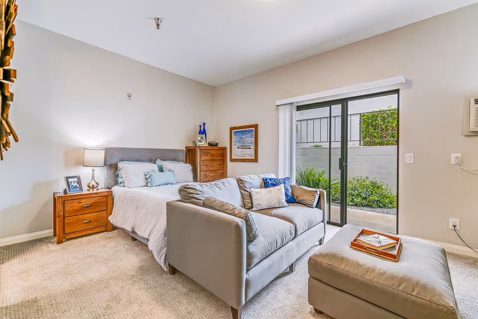 A bright and cozy bedroom in Rancho Vista Senior Living featuring a bed with white bedding and multiple pillows, a wooden nightstand with a lamp and framed photo, a wooden dresser with decorative items, a gray loveseat with cushions, and a matching ottoman with a tray. Sliding glass doors open to a small outdoor garden area.