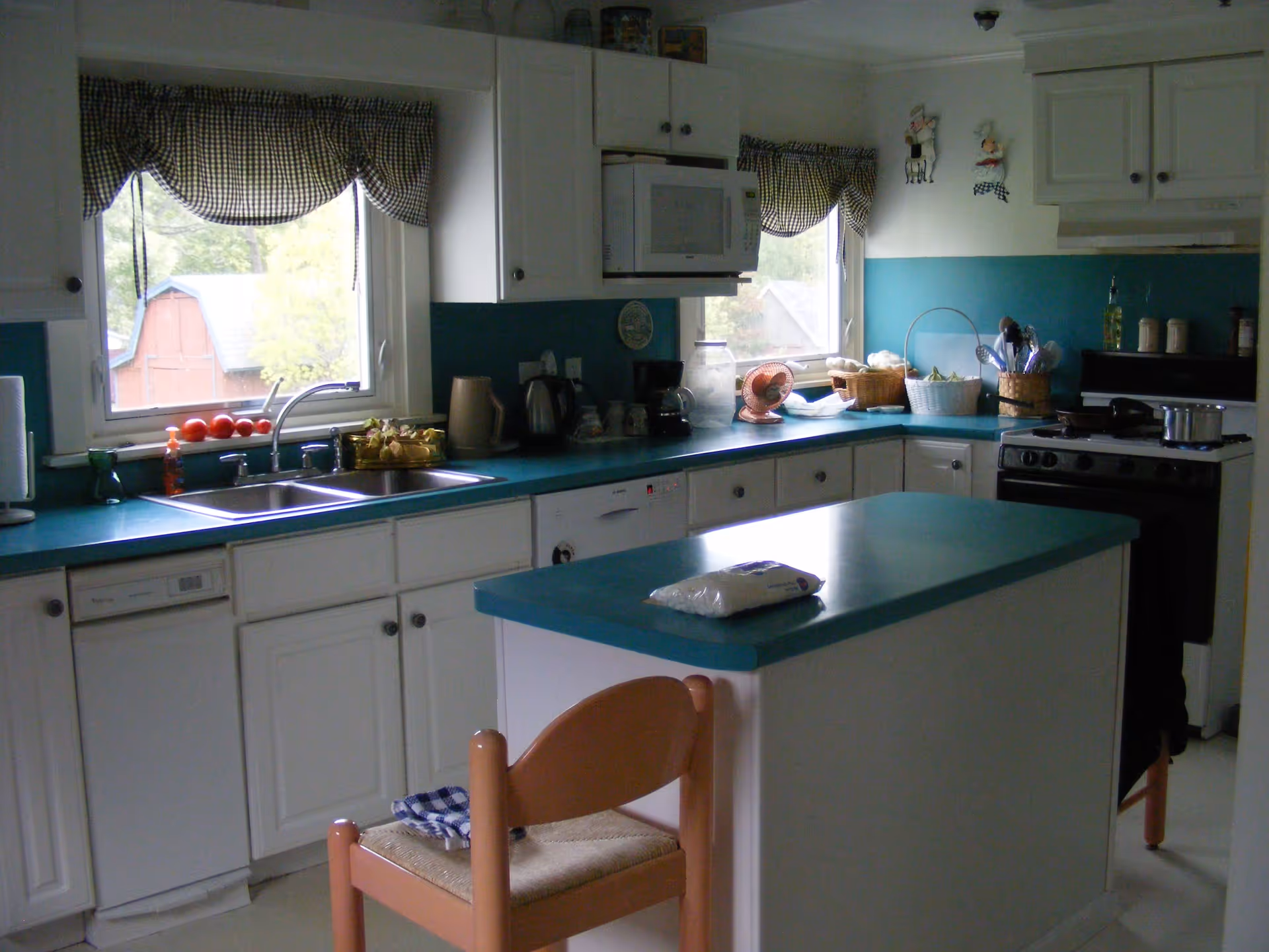 A kitchen with white cabinets and teal countertops. There is a double sink under a window with checkered curtains, a microwave above the counter, a stove with pots, and various kitchen items like a coffee maker, baskets, and utensils. A small island with a teal countertop and a wooden chair with a woven seat are in the foreground.