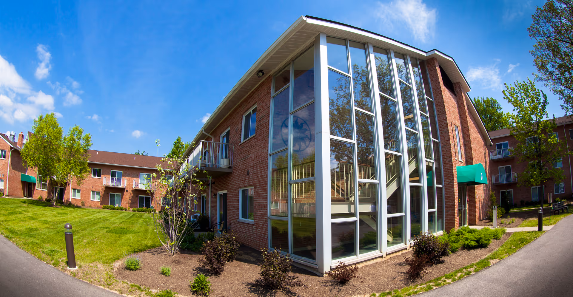 Brick senior living building exterior with a large multi-story glass stairwell, green lawn, and walkways under a blue sky.