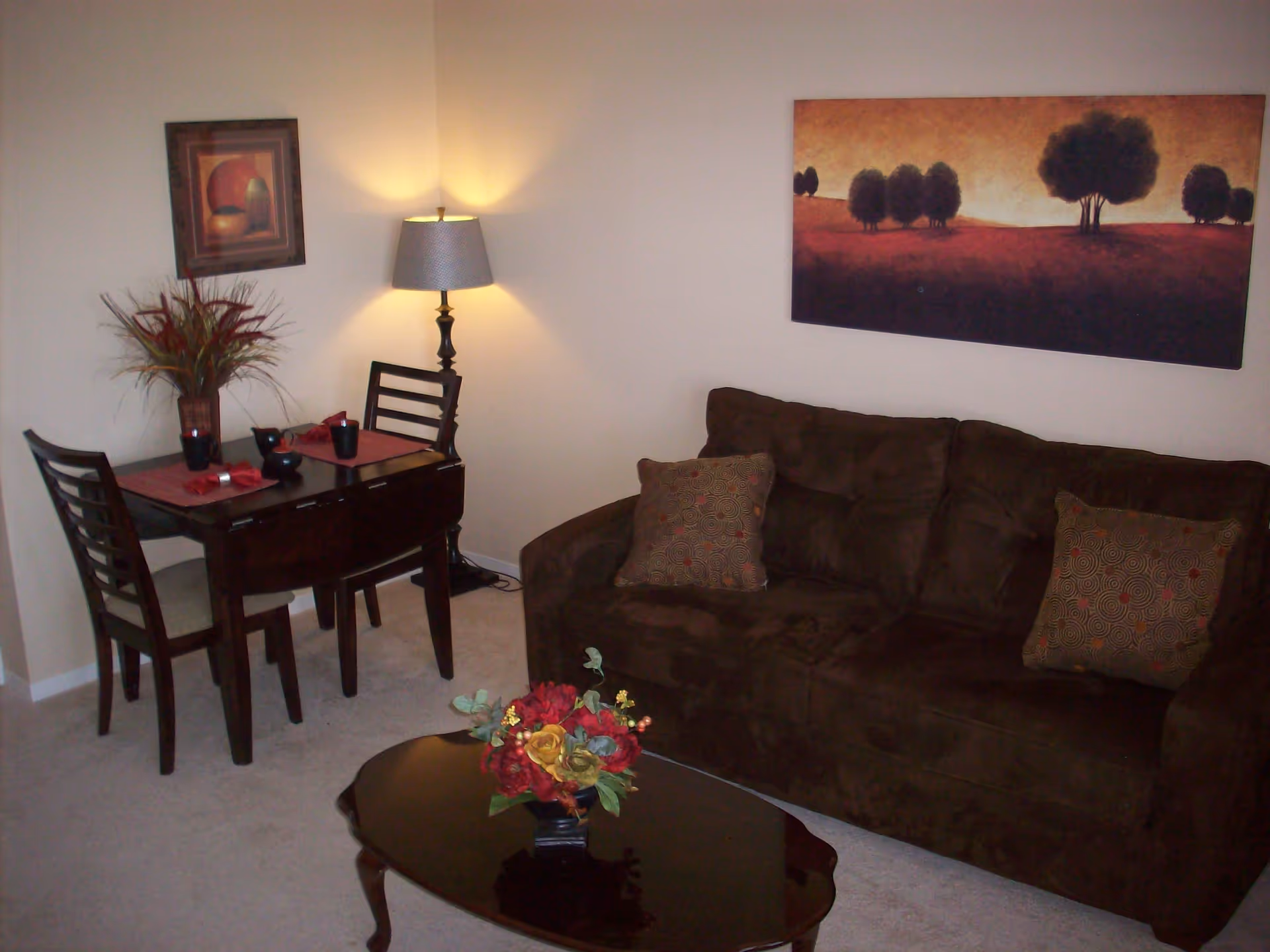 A cozy living room area with a dark brown sofa adorned with two patterned cushions, a wooden coffee table with a floral arrangement, and a small dining table set for two with placemats, napkins, and cups. A floor lamp illuminates the corner, and two framed artworks hang on the beige walls.