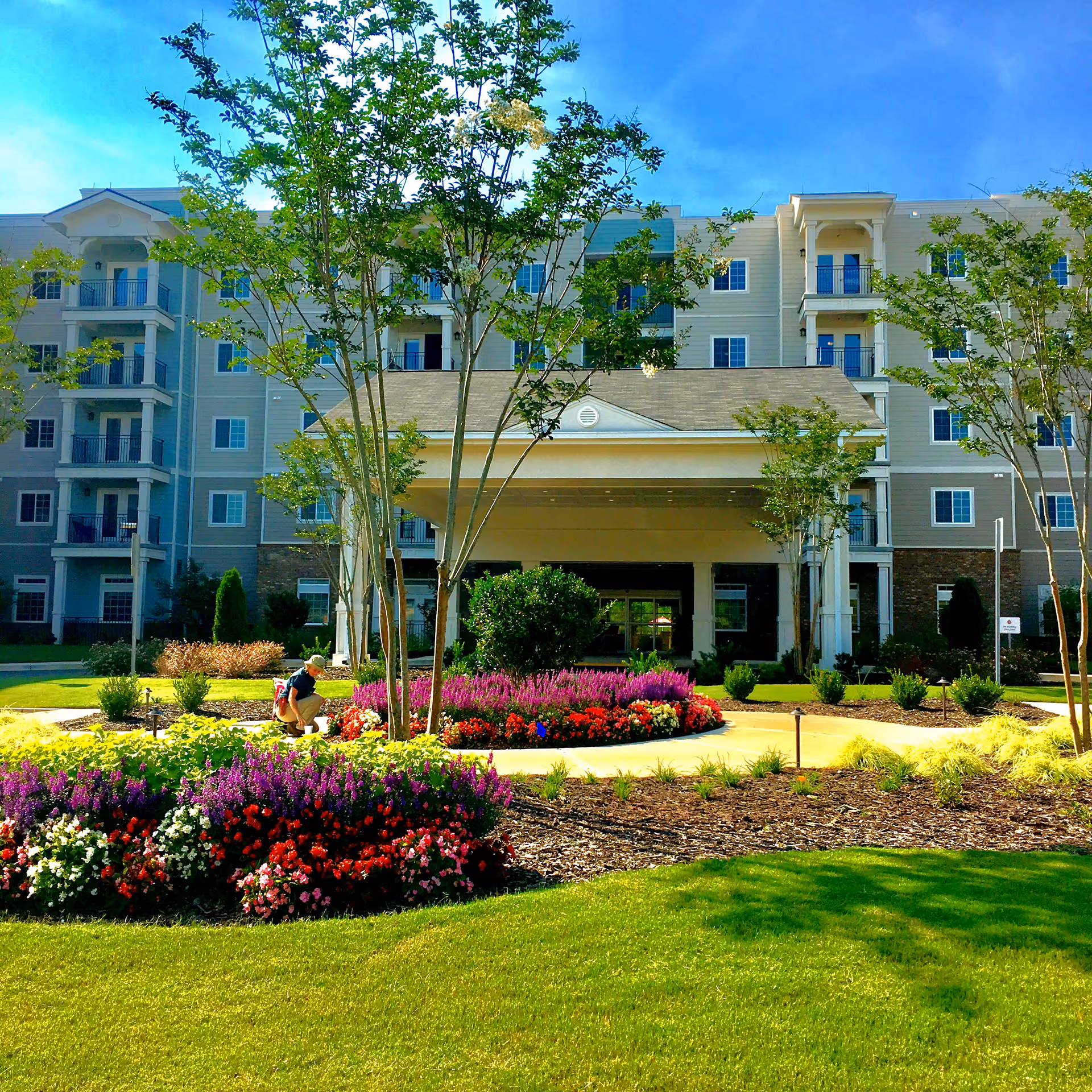 Front entrance of a multi-story senior living building with a porte-cochere, trees and colorful landscaped flowerbeds.