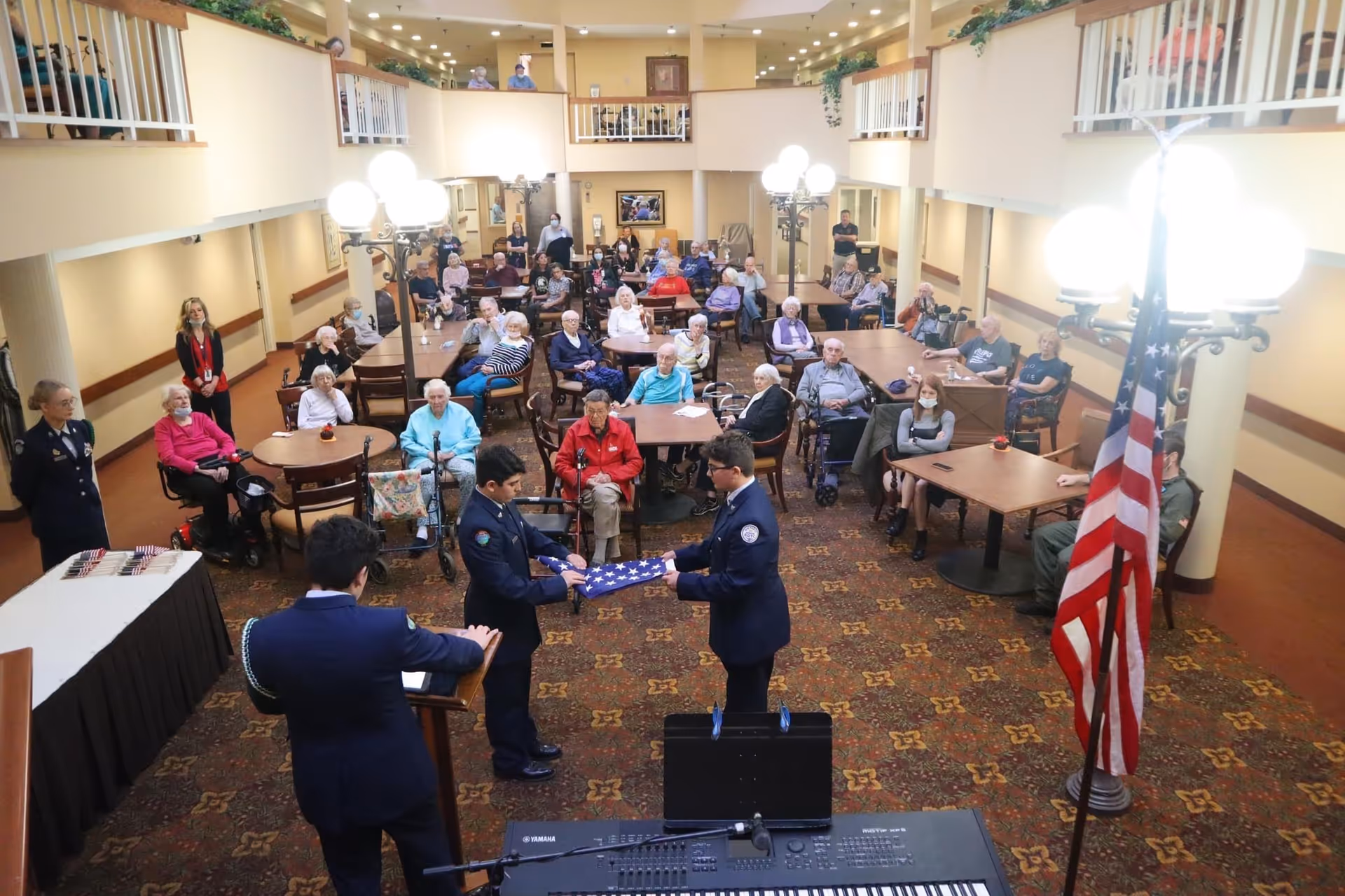Residents seated at tables in a senior living common room watching uniformed personnel perform a flag-folding ceremony.