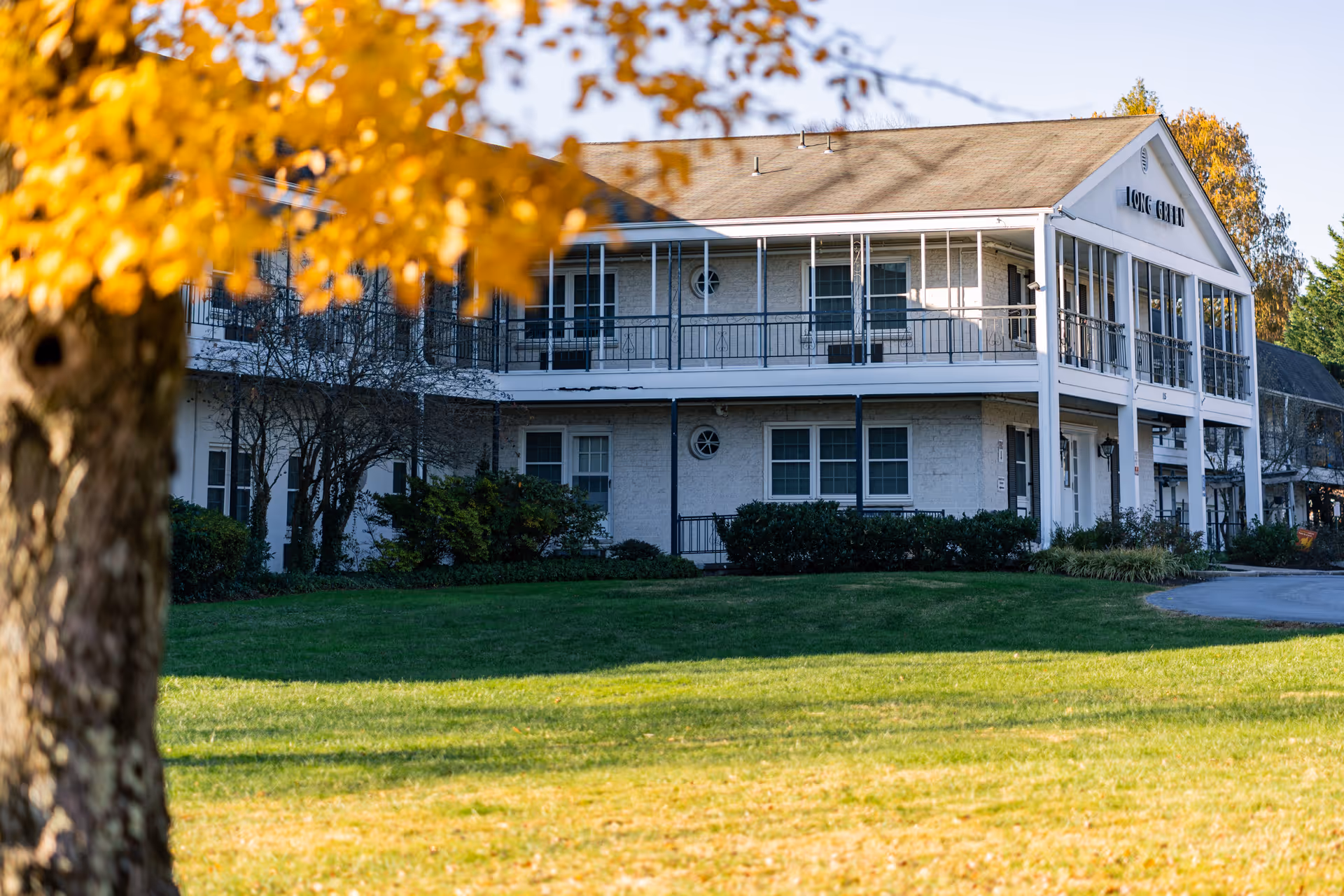 Exterior view of a two-story building with white walls and a balcony, surrounded by green grass and trees with autumn-colored leaves in the foreground.