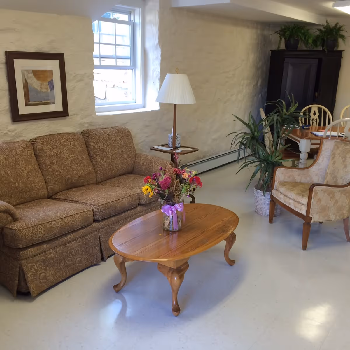A cozy living room area with a patterned brown sofa, a wooden coffee table with a vase of colorful flowers, a beige upholstered armchair, a floor lamp, and a window letting in natural light. In the background, there is a dining table with chairs and a dark wooden cabinet with plants on top.