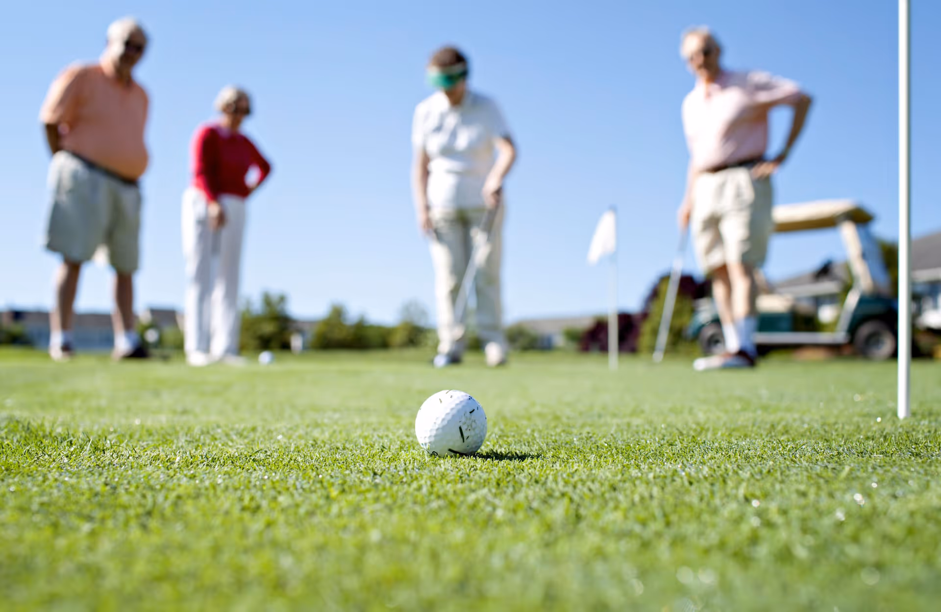 A close-up of a golf ball on the grass with four elderly people in the background playing golf on a sunny day. A golf cart is visible to the right.