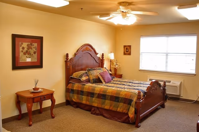 A cozy bedroom with a wooden bed frame featuring intricate carvings, a colorful plaid bedspread, and multiple pillows. There is a wooden nightstand with a lamp and decorative items next to the bed, a framed floral artwork on the wall, and a window with blinds letting in natural light. The room has beige walls and carpeted flooring.