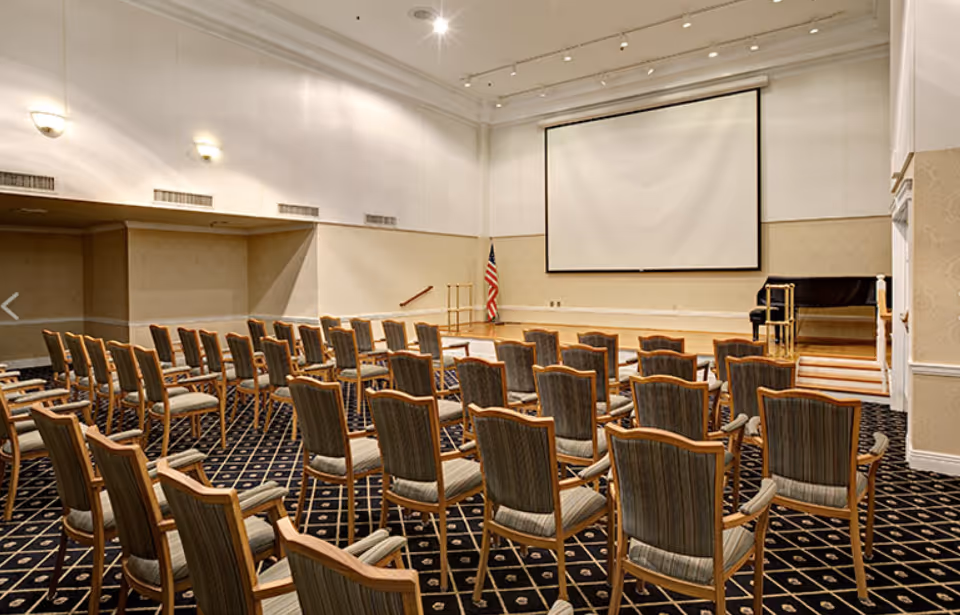 A conference or presentation room with rows of wooden chairs with padded seats and backs arranged facing a stage. The stage has a large projection screen, an American flag, and a piano. The room has patterned carpet and neutral-colored walls with wall-mounted lights.