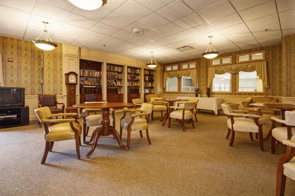 A cozy common area with multiple round wooden tables surrounded by cushioned armchairs. The room features patterned wallpaper, a carpeted floor, and three large windows with valances. There are built-in bookshelves filled with books along one wall, a grandfather clock, and a television on a stand in the corner. Soft ceiling lights illuminate the space.
