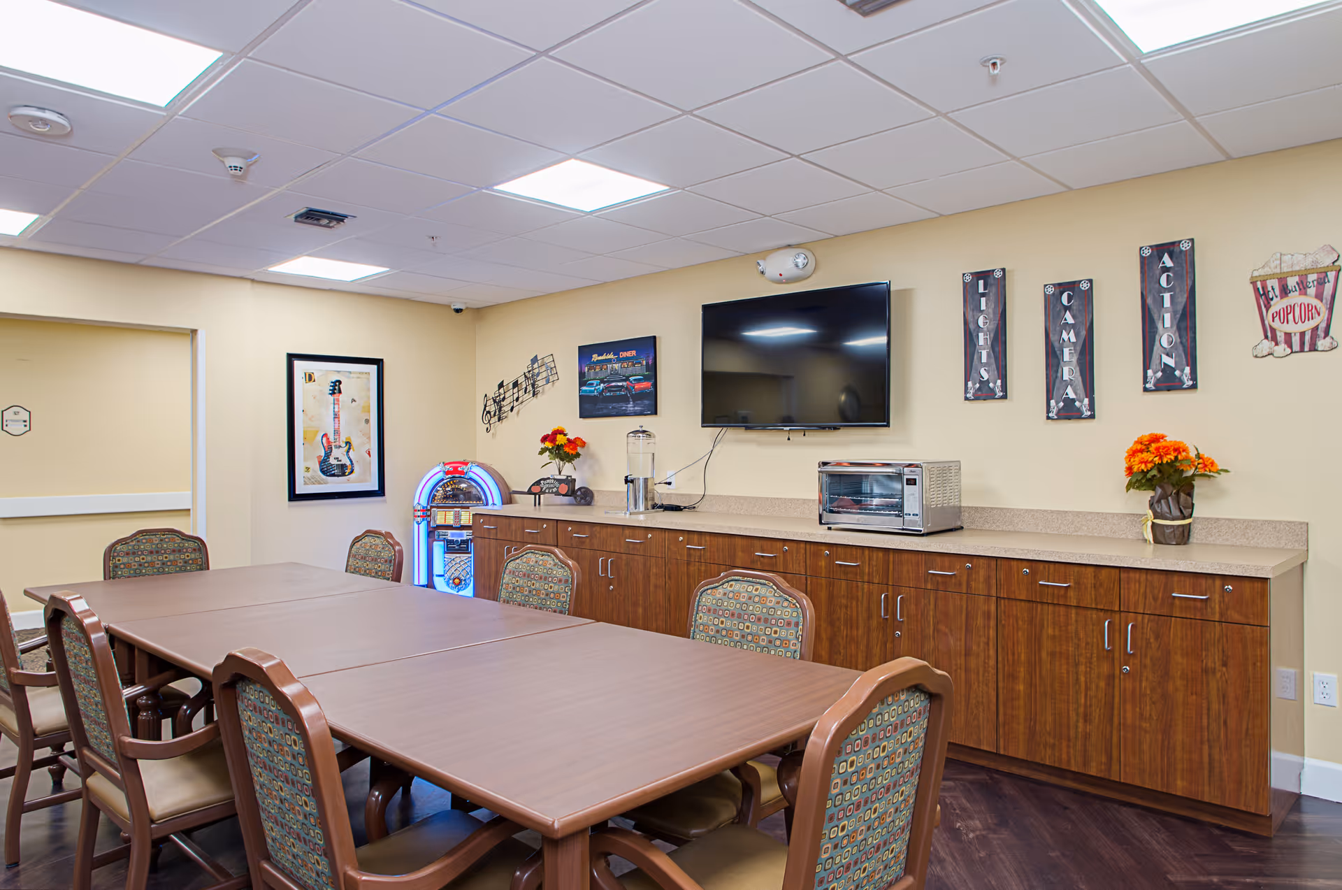A meeting or activity room with a large wooden table surrounded by chairs with patterned upholstery. On the wall, there is a flat-screen TV, a toaster oven, a water dispenser, and decorative wall art including musical notes, a guitar picture, and signs that say 'Lights', 'Camera', 'Action', and a popcorn bucket. The room has a drop ceiling with fluorescent lighting and wood flooring.
