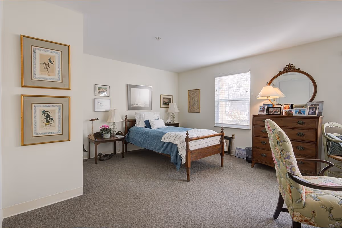 A cozy bedroom with a wooden bed covered in blue and white bedding, two bedside tables with lamps, framed artwork on the walls, a window letting in natural light, a wooden dresser with a mirror and several framed photos, and a floral upholstered chair.
