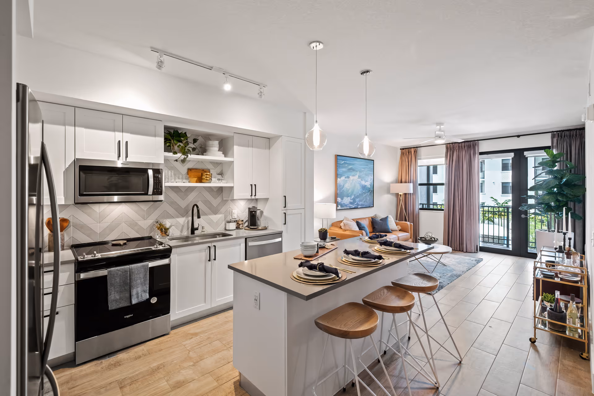 Open-plan modern kitchen with an island and bar stools opening into a living area with a sofa and large windows.