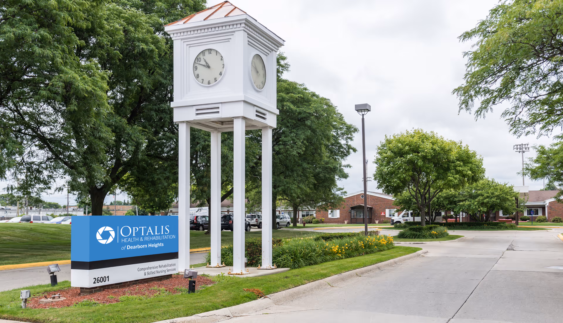 Outdoor view of Optalis Health & Rehabilitation of Dearborn Heights facility entrance with a large white clock tower, a blue and white facility sign, green trees, and a driveway leading into the property.