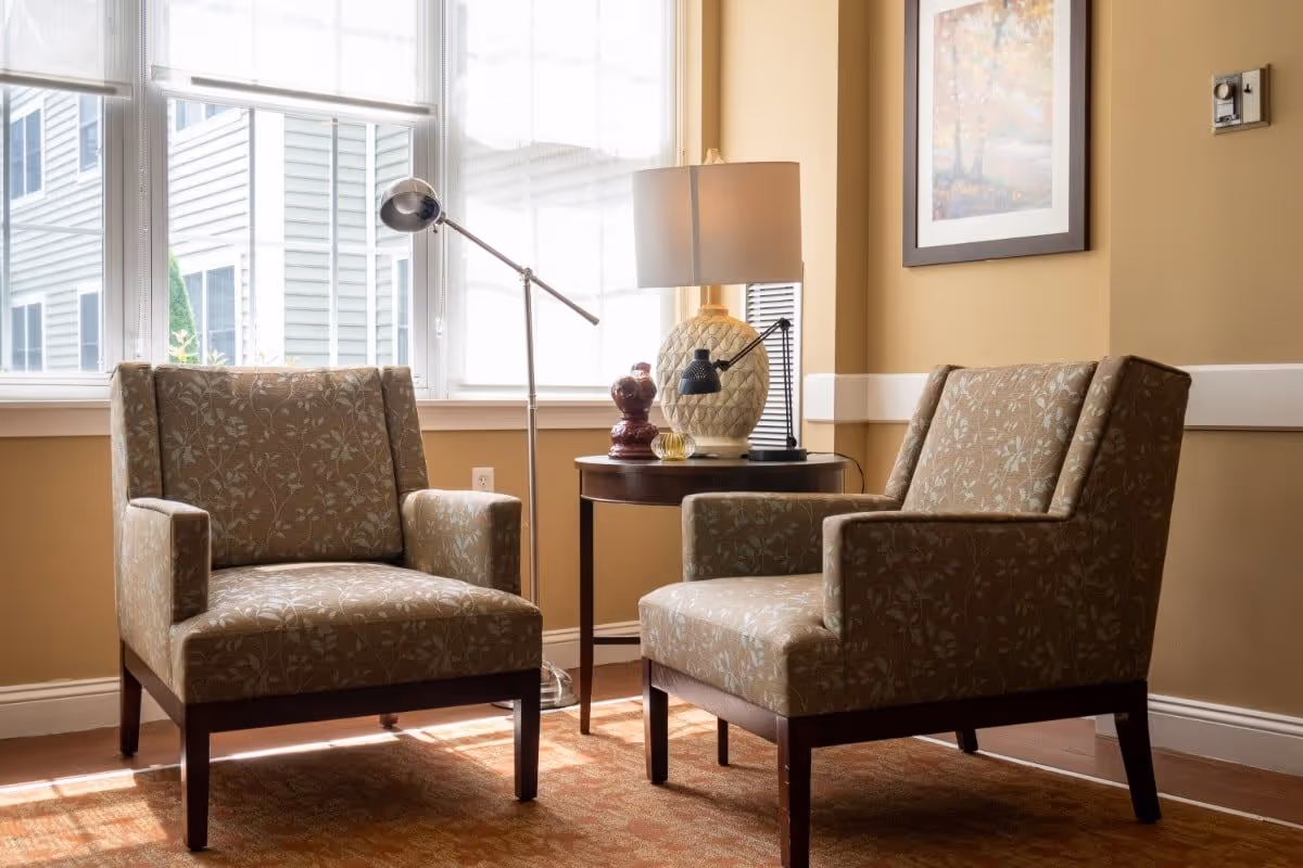 A cozy seating area with two patterned armchairs facing each other, a round wooden side table between them holding a decorative lamp and small items, next to a large window with white blinds letting in natural light. A framed artwork hangs on the beige wall.