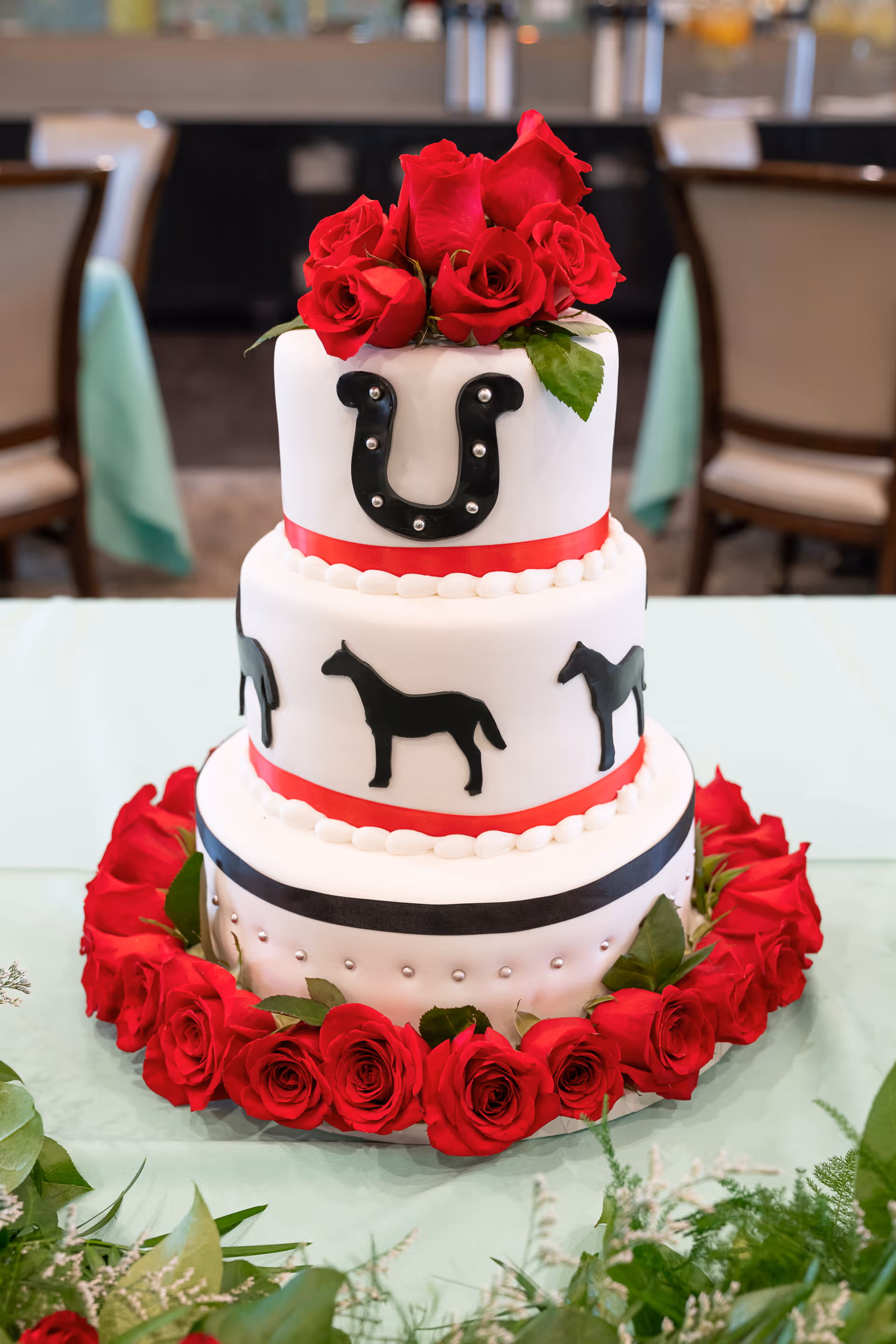 A three-tier white cake decorated with black horse silhouettes and a black horseshoe on the top tier. The cake is adorned with red ribbons and surrounded by red roses at the base and on top. The background shows a dining area with chairs and tables.