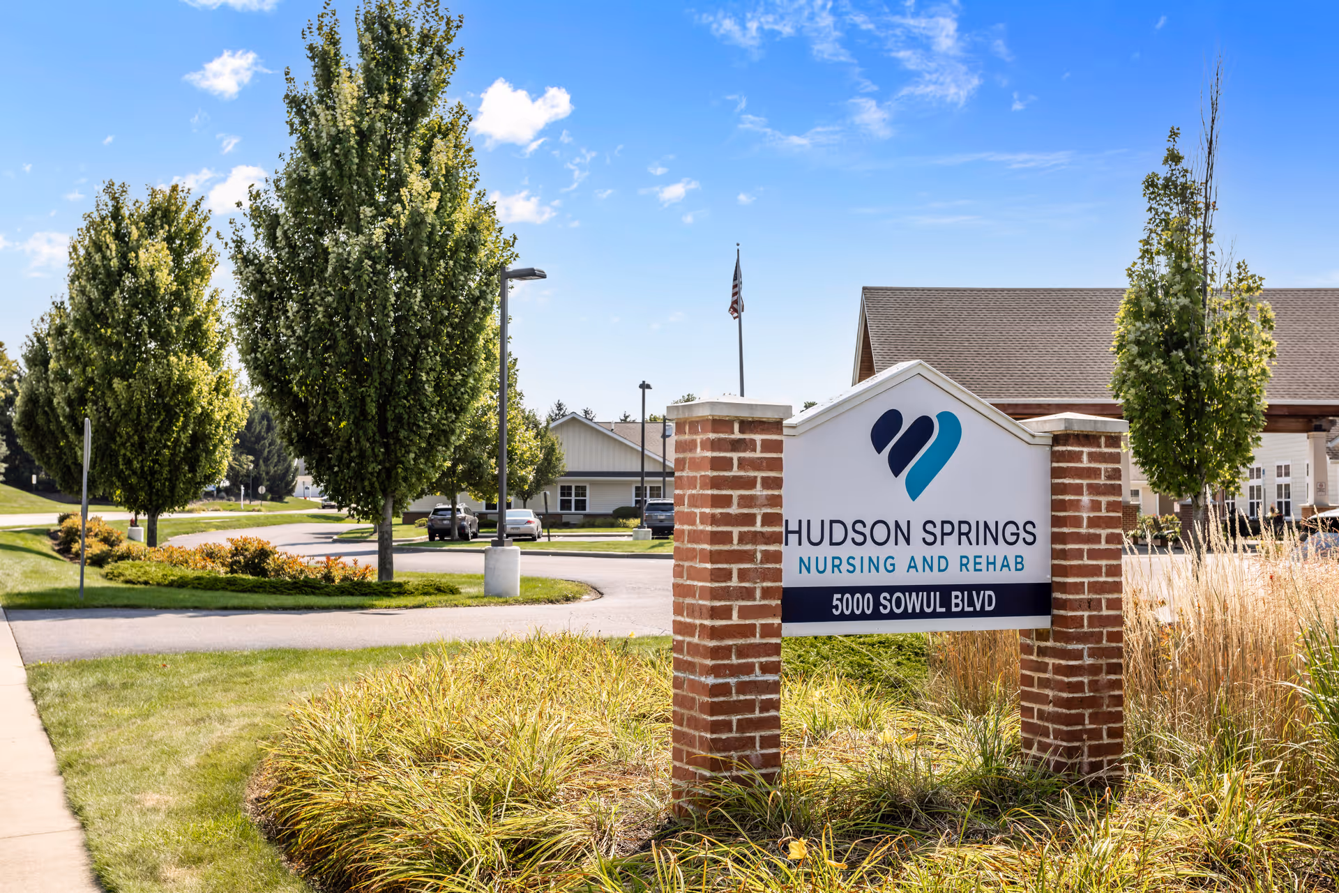 Outdoor view of Hudson Springs Nursing and Rehab facility sign with address 5000 Sowul Blvd, surrounded by grass, trees, and a clear blue sky.