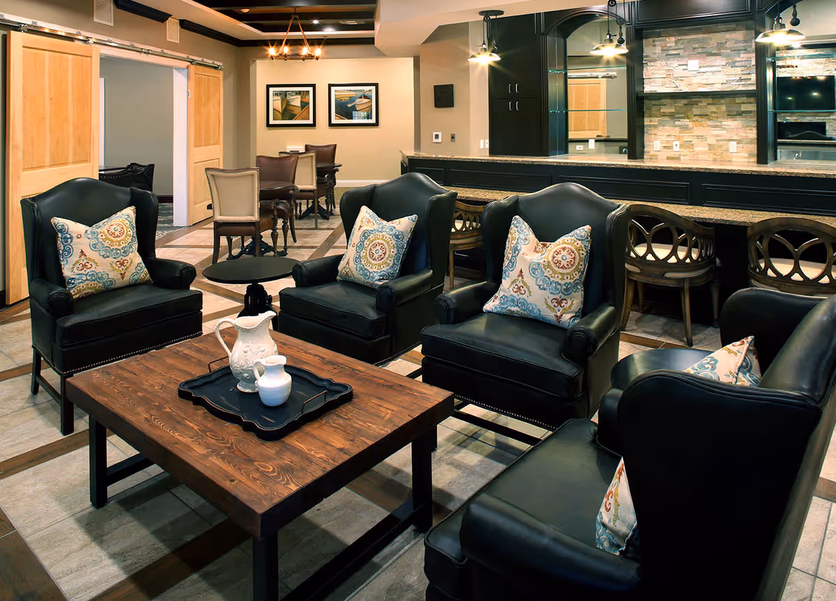A cozy living room area with four black leather armchairs, each adorned with a decorative patterned pillow. In the center is a wooden coffee table with a black tray holding a white pitcher and a small white container. Behind the seating area is a long counter with bar stools and a stone accent wall with a mounted TV. The room also features a dining area with tables and chairs, wooden sliding doors, and framed pictures on the wall.