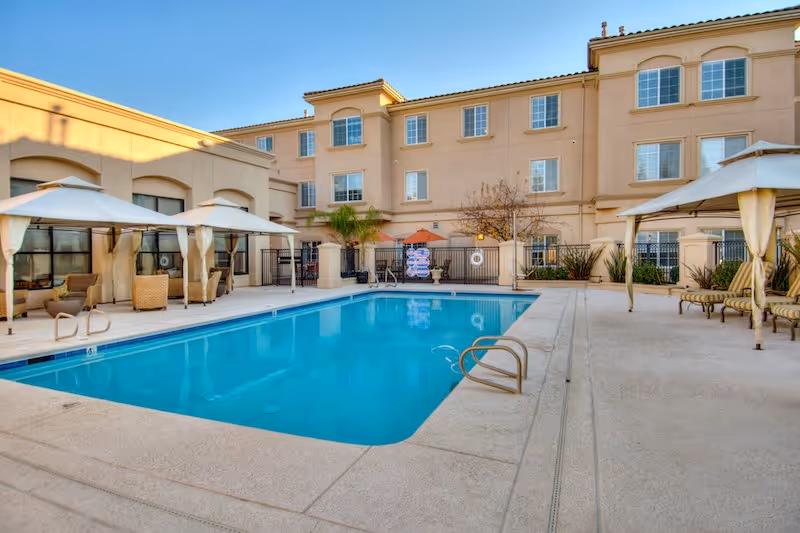 Outdoor swimming pool area at a senior living facility with lounge chairs, umbrellas, and a three-story beige building in the background under a clear blue sky.