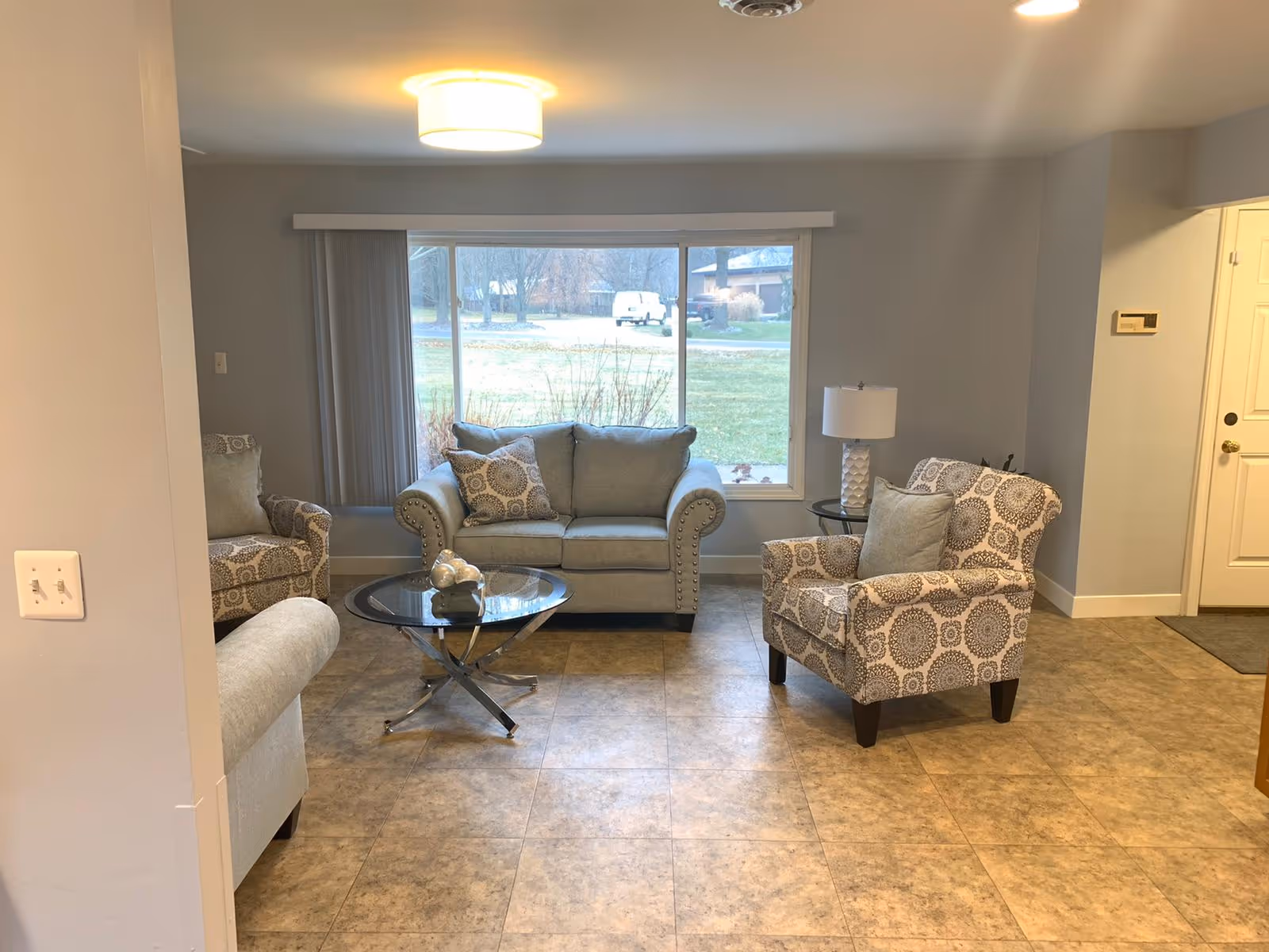 A cozy living room with a large window showing an outdoor view. The room features a gray loveseat with two patterned pillows, two patterned armchairs with cushions, a glass coffee table with decorative items, a side table with a lamp, and tiled flooring. The walls are painted light gray and there is a ceiling light fixture.