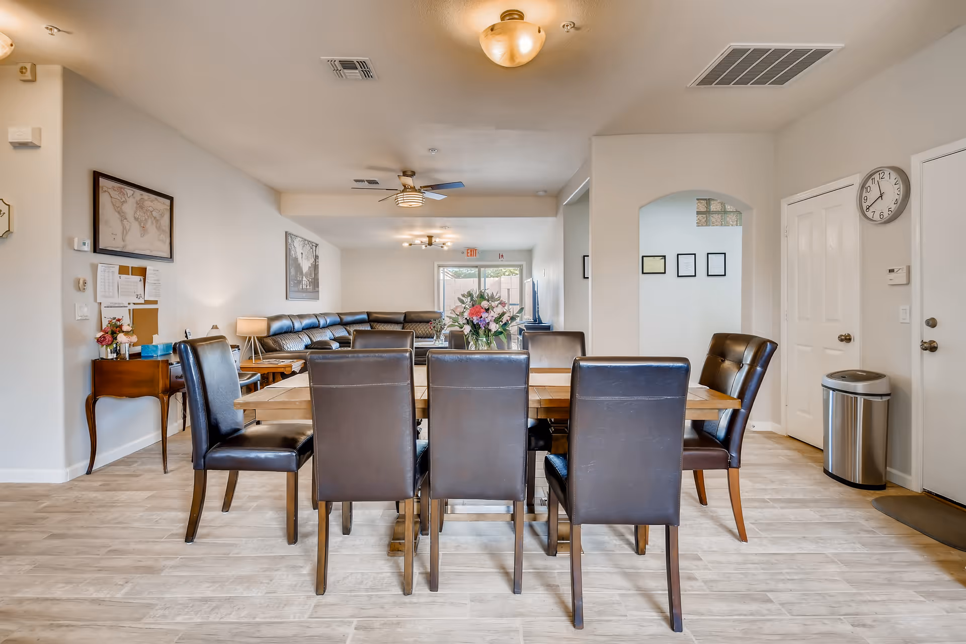 A bright and spacious dining and living area in an assisted living facility. The foreground features a wooden dining table surrounded by eight dark leather chairs. In the background, there is a large sectional sofa with a ceiling fan above it. The walls are decorated with framed pictures and a world map. A clock and a trash can are visible near a white door on the right side. The floor is covered with light-colored wood-like tiles.
