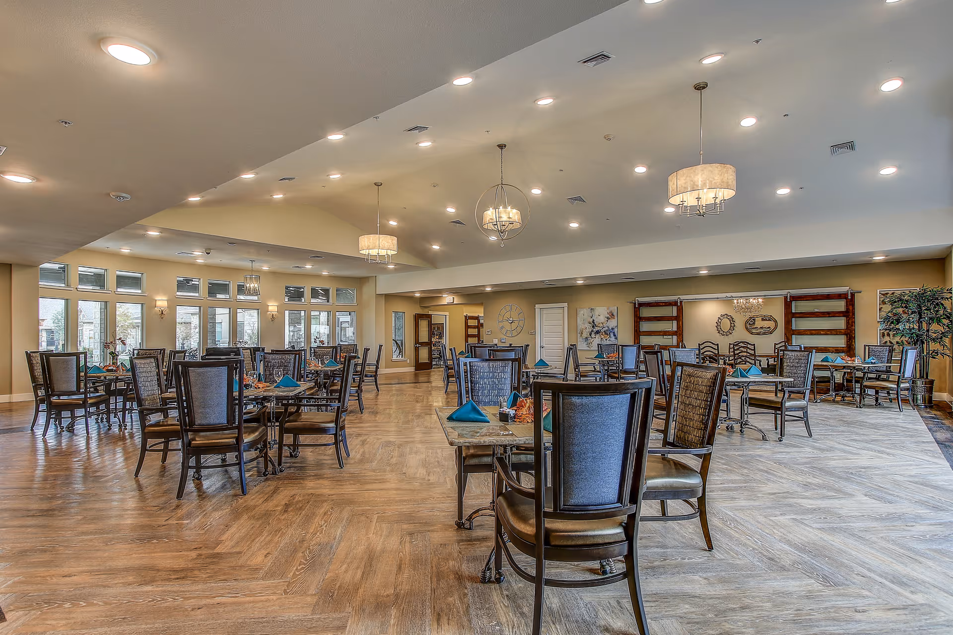 A spacious dining room with multiple tables and chairs arranged neatly. Each table has place settings with blue napkins. The room features large windows allowing natural light to enter, modern ceiling lights, and decorative wall art. The floor has a wood pattern, and there are plants and shelves along the walls.